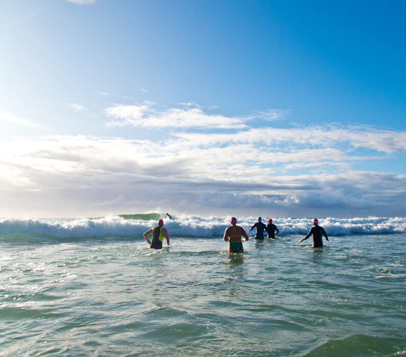A group of swimmers, including blind athlete, Gerrard Gosens, walk out through the waves at a Gold Coast beach on a nice blue sky day. Gosens is wearing a black wetsuit and is tethered to a friend who will guide him through the waves and in the open water.
